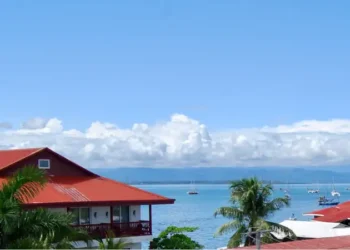 Panoramic view of Bocas del Toro Bay with red-roofed waterfront homes, palm trees, and sailboats anchored on calm tropical waters under a bright blue sky.