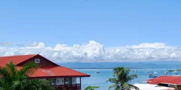 Panoramic view of Bocas del Toro Bay with red-roofed waterfront homes, palm trees, and sailboats anchored on calm tropical waters under a bright blue sky.