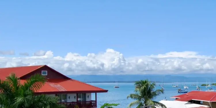 Panoramic view of Bocas del Toro Bay with red-roofed waterfront homes, palm trees, and sailboats anchored on calm tropical waters under a bright blue sky.