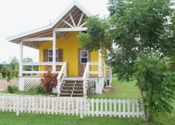 Bright yellow cottage with a front porch, white picket fence, and lush green yard surrounded by trees.