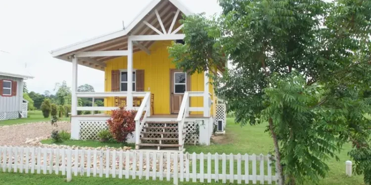 Bright yellow cottage with a front porch, white picket fence, and lush green yard surrounded by trees.