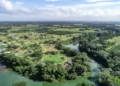 An aerial view of a lush rural area with a winding river, dense green trees, scattered homes, and farmland stretching into the distance under a partly cloudy sky.