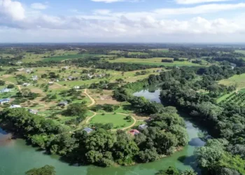 An aerial view of a lush rural area with a winding river, dense green trees, scattered homes, and farmland stretching into the distance under a partly cloudy sky.