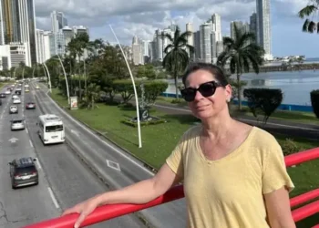Kathleen stands on a red pedestrian bridge overlooking a busy coastal road, with palm trees, the ocean, and Panama City’s modern skyline behind her.