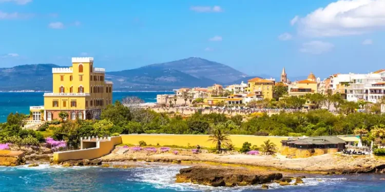 Mediterranean seacoast in Alghero city, Sardinia, Italy. Spring flowers and trees on foreground, colourful buildings of Alghero old city center on background
