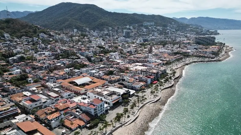 aerial view of a coastal city in Mexico shows a dense town with red-roofed buildings along a palm-lined waterfront, backed by green mountains and bordered by the ocean.