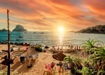 Picturesque view of Cala d'Hort tropical Beach, people hangout in beautiful beach with Es Vedra rock view during magnificent vibrant sunset glowing sun. Balearic Islands, Spain, Espana. Ibiza