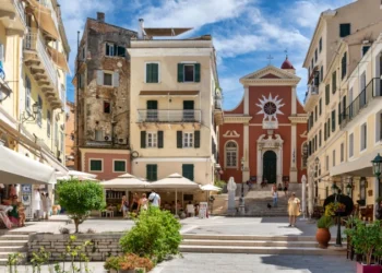 A vibrant scene of daily life in Corfu, Greece, showing people in Mitropolis Square with the red facade of the Metropolitan Cathedral of Panagia Spiliotissa in the background.