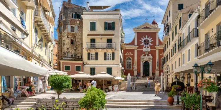 A vibrant scene of daily life in Corfu, Greece, showing people in Mitropolis Square with the red facade of the Metropolitan Cathedral of Panagia Spiliotissa in the background.