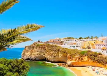 A sunny coastal scene in the Algarve, showing colorful seaside houses perched atop golden limestone cliffs above a small sandy beach.