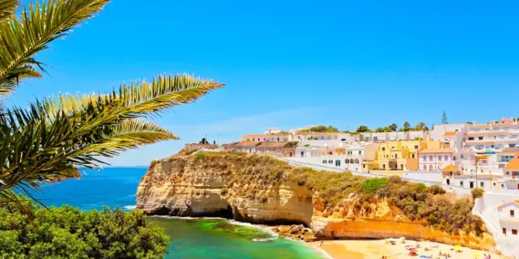 A sunny coastal scene in the Algarve, showing colorful seaside houses perched atop golden limestone cliffs above a small sandy beach.