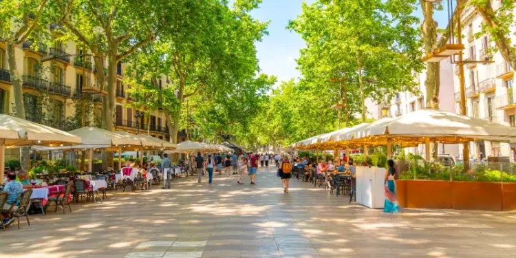 Tourists walk past sidewalk cafes on the famous La Rambla street through the historic Mediterranean city of Barcelona, Catalonia, Spain.