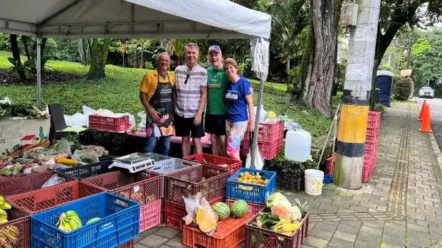 Four people pose together under a canopy in a park, surrounded by crates of fruits and vegetables at an outdoor market stall.