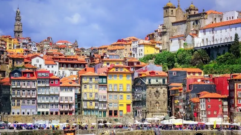 Colorful riverside buildings line the waterfront in Porto, with historic churches and terracotta rooftops rising up the hillside, creating a vibrant and iconic cityscape along the Douro River.