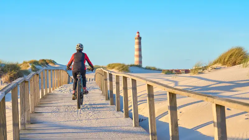 senior woman cycling during moody golden hour at the beach of the atlantic coast below Barra Lighthouse near Aveiro, Portugal