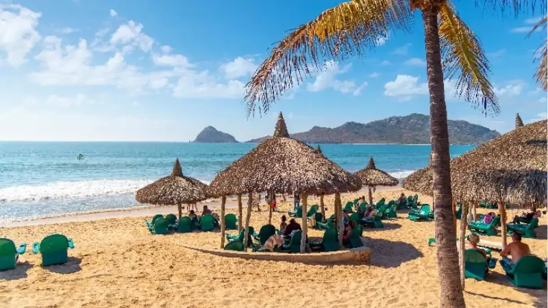 A sunny beach scene with thatched-roof huts, lounge chairs, and people relaxing by the ocean with mountains in the distance.