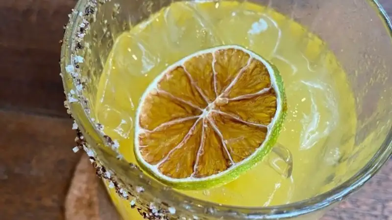 Refreshing citrus margarita, likely lime or lemon, with a salted rim, ice, and a dried lime slice garnish in a textured glass.