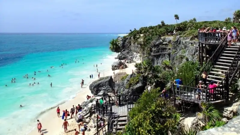 Crowded tropical beach with turquoise water, rocky cliffs, and wooden stairways overlooking the shoreline.