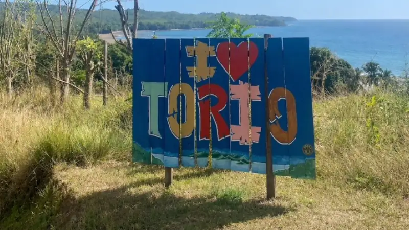 A colorful “I love Torio” sign stands on a grassy hill overlooking the ocean, with a rugged coastline, trees, and clear blue sky in the background.