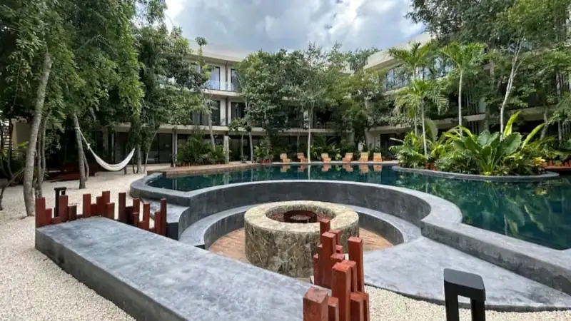 Lush tropical courtyard with a modern pool, surrounded by greenery, palm trees, and multi-level resort buildings under a partly cloudy sky.