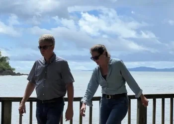 Lief and Kathleen walk along a wooden railing by the ocean under a cloudy sky, with a small tree-covered island in the background.