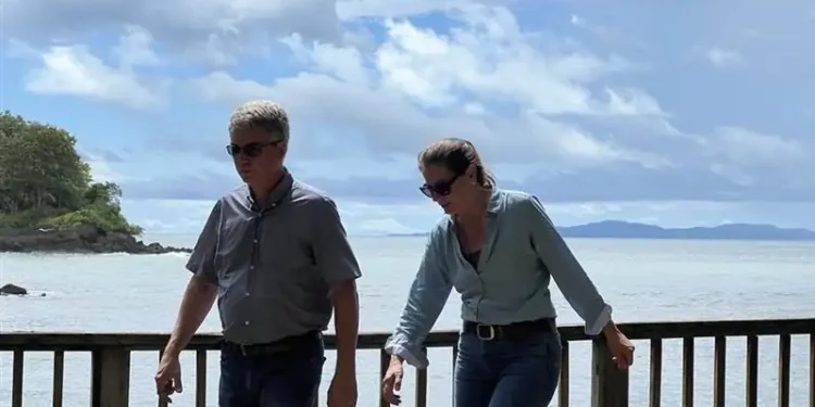 Lief and Kathleen walk along a wooden railing by the ocean under a cloudy sky, with a small tree-covered island in the background.