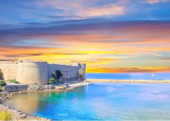Historic seaside fortress in North Cyprus overlooking calm turquoise waters at sunset, with soft golden and pastel skies reflecting across the coastline.