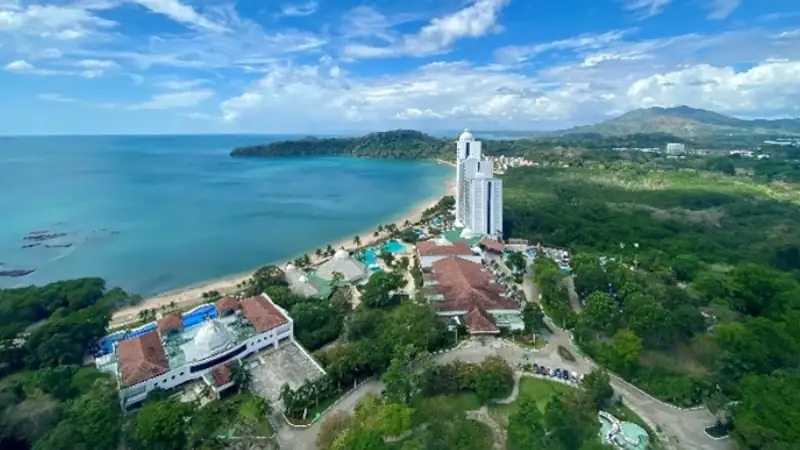 Aerial view of a tropical beachfront resort with tall white buildings, sandy shoreline, and turquoise ocean surrounded by lush green hills.