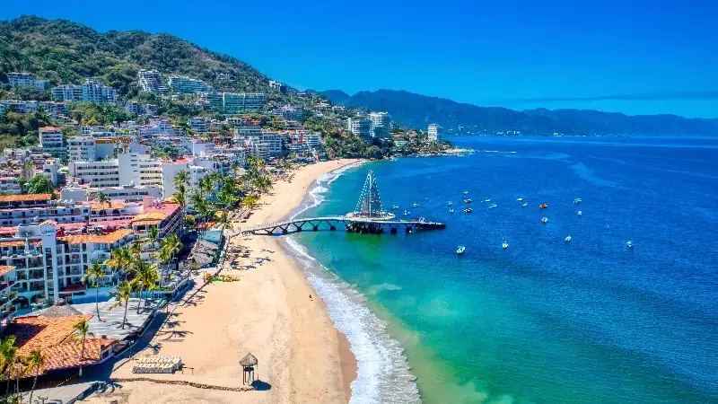 A sunlit coastal view of Puerto Vallarta featuring a sandy beach, palm-lined promenade, and the iconic pier extending into calm turquoise waters, with mountains rising in the background.