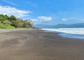 Wide black-sand beach with gentle waves, lush tropical greenery, and distant mountains under a bright blue sky.