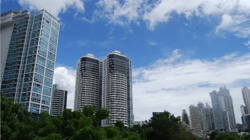 Modern high-rise buildings and residential towers rising above greenery under a bright blue sky in Panama City.