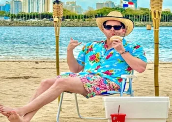 Mayer, the mayor of Panama City, relaxes on the beach in colorful tropical clothing while enjoying a drink, with the Panama City skyline visible in the background.
