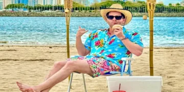 Mayer, the mayor of Panama City, relaxes on the beach in colorful tropical clothing while enjoying a drink, with the Panama City skyline visible in the background.