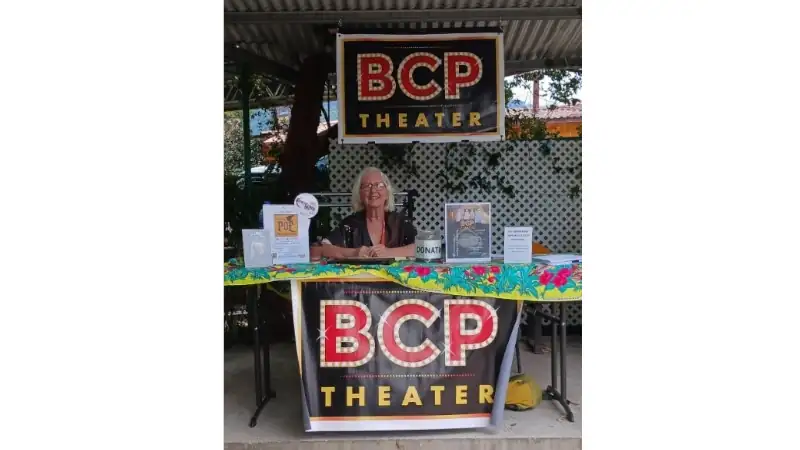 Woman seated at a BCP Theater booth during a local community event with theater signs and donation materials on display.