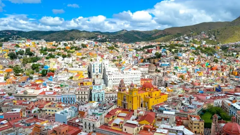 A vibrant cityscape with colorful buildings spread across rolling hills under a bright blue sky.