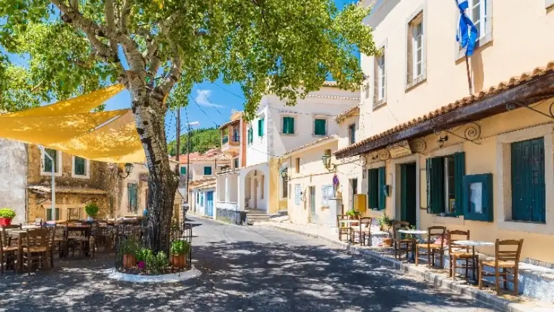 Quiet village street lined with traditional Mediterranean houses, outdoor café seating, and a large shade tree beneath a clear blue sky.