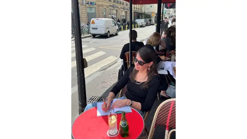 Kathleen relaxing at a Paris sidewalk café while writing in a notebook and enjoying a quiet afternoon drink.