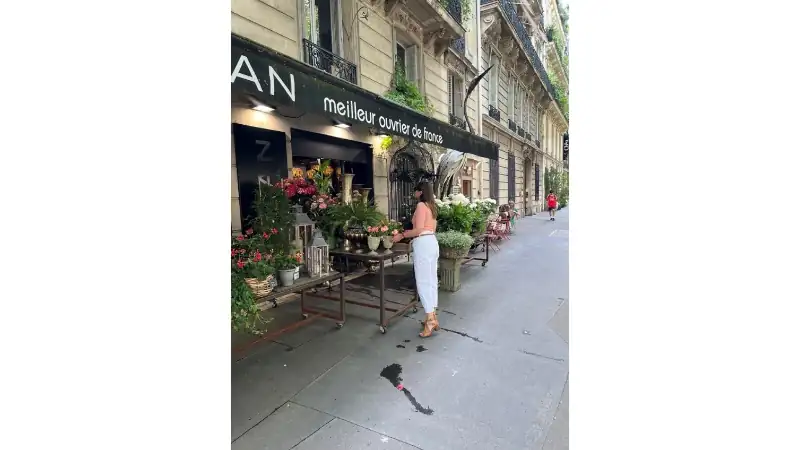 Kathleen strolling past a charming flower shop on a classic Paris street lined with elegant buildings and outdoor seating.