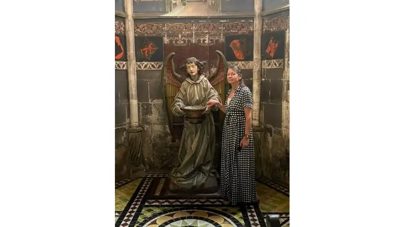 Kathleen standing beside an angel statue inside a historic chapel with ornate medieval architecture and patterned tile flooring.