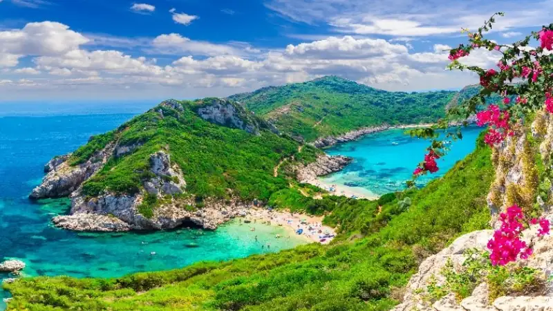 Panoramic view of a turquoise Mediterranean cove surrounded by lush green hills, rocky cliffs, and vibrant pink bougainvillea under a bright blue sky.