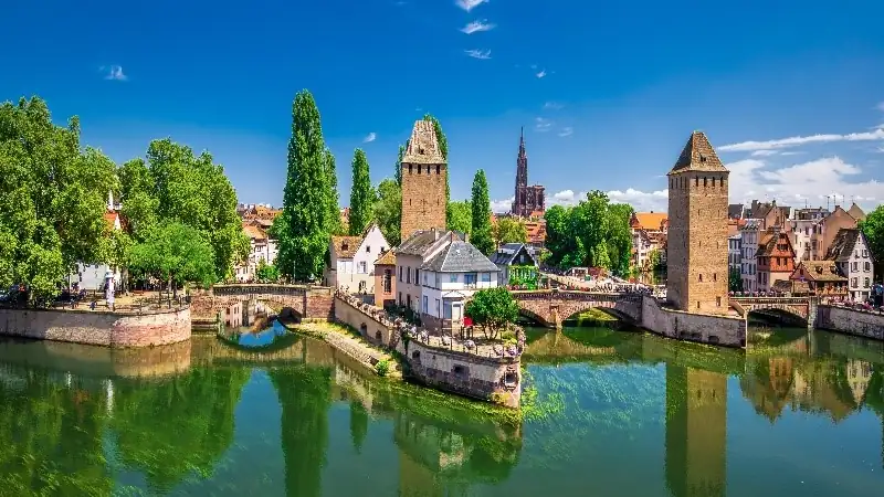 Scenic view of Strasbourg’s historic canals, stone towers, and half-timbered buildings under a bright blue sky.