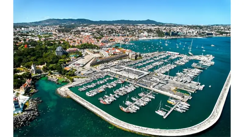An aerial view of a modern marina filled with yachts, surrounded by a vibrant coastal city, clear blue waters, and a scenic waterfront backed by rolling hills.