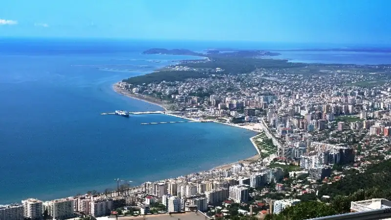 An aerial view of a vibrant coastal city featuring a sweeping shoreline, modern buildings, a scenic bay, and the deep blue sea stretching toward distant islands.