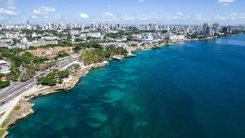 A panoramic aerial view of a vibrant coastal city with turquoise waters, a scenic shoreline, and a modern skyline under a bright blue sky with scattered clouds.