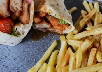 Crispy chicken wrap filled with fresh lettuce, pickled onions, roasted tomatoes, and sauce, served with seasoned French fries on a ceramic plate.