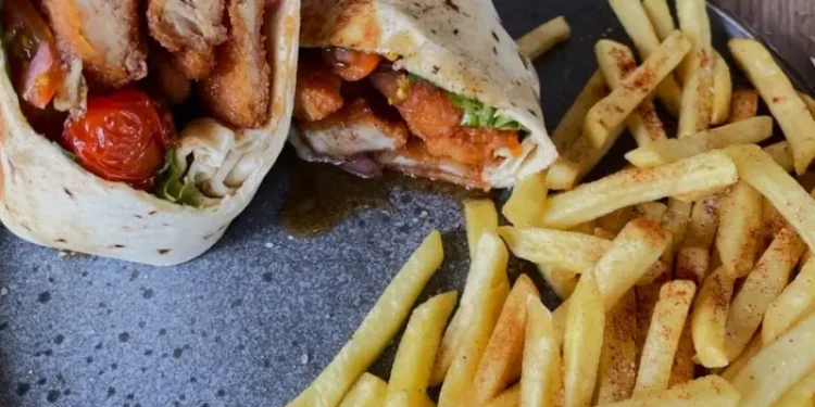 Crispy chicken wrap filled with fresh lettuce, pickled onions, roasted tomatoes, and sauce, served with seasoned French fries on a ceramic plate.