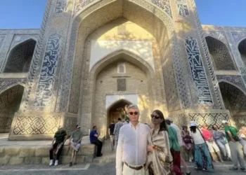 Lief and Kathleen pose in front of a grand historic Islamic landmark in Bukhara, Uzbekistan, surrounded by intricate blue-tiled architecture and fellow travelers on a sunny afternoon.