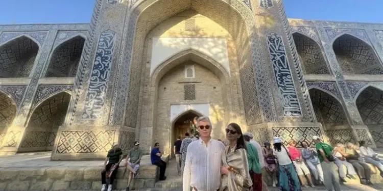 Lief and Kathleen pose in front of a grand historic Islamic landmark in Bukhara, Uzbekistan, surrounded by intricate blue-tiled architecture and fellow travelers on a sunny afternoon.