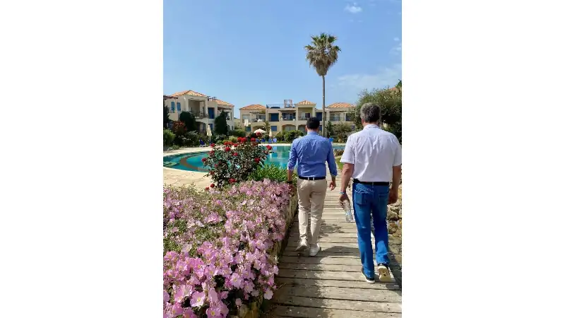 Lief walks alongside another man on a wooden path beside a resort pool, surrounded by blooming flowers, palm trees, and Mediterranean-style villas under a clear blue sky.