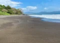 A wide, tranquil black sand beach bordered by lush tropical forest, with gentle waves, distant mountains, and a bright blue sky dotted with soft clouds.